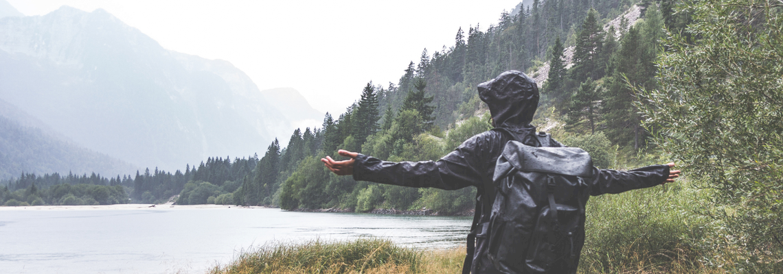Eine Person in einer Regenjacke steht mit einem Rucksack auf dem Rücken in der Natur.